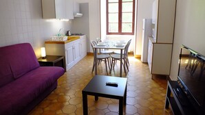 A compact living space with a purple sofa, a dining table with chairs, a kitchen area with a sink and cabinets, and a TV. - Studio in the heart of the village (Saint-Jean-du-Bruel)
