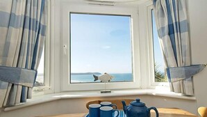 A room with a bay window overlooking the sea, a table with a blue and white striped tablecloth, two blue mugs, and a blue teapot.