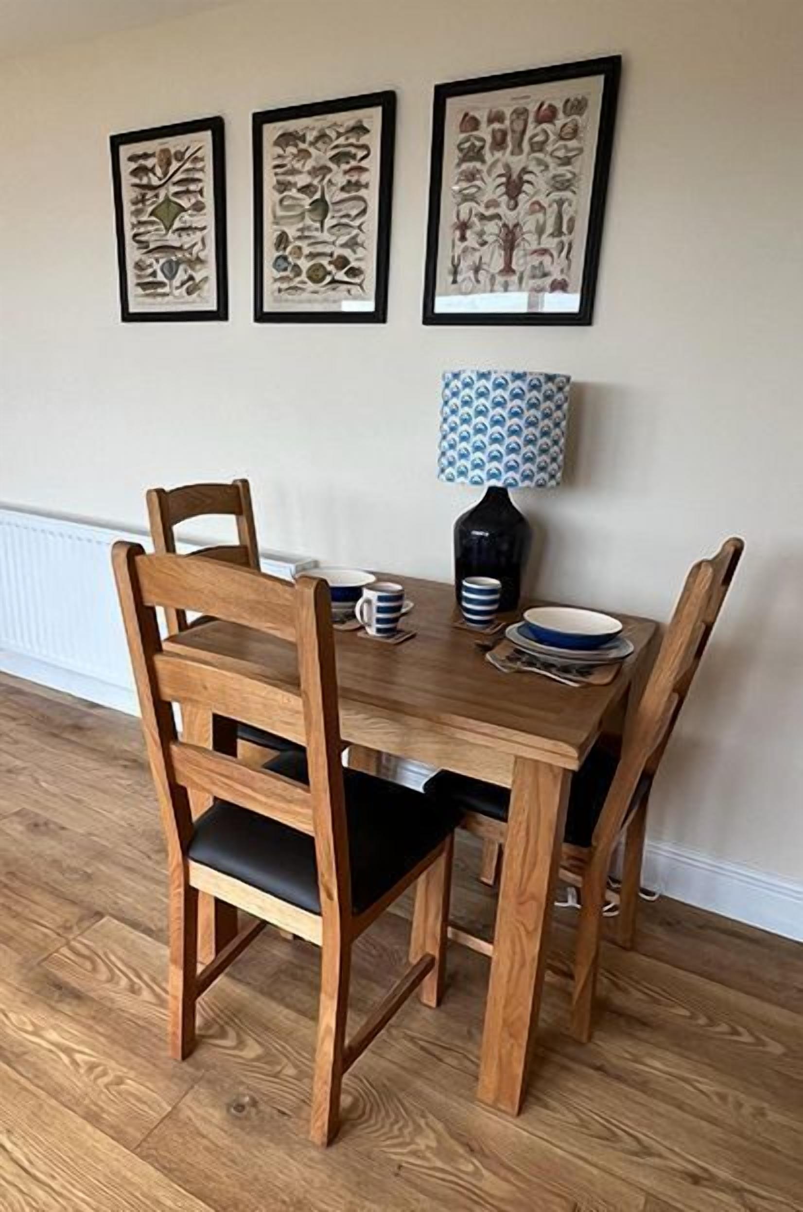 A dining table with four wooden chairs, a vase with a patterned lampshade, and three framed botanical prints on the wall.