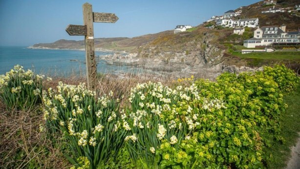 A coastal path signpost with a view of houses on a hillside and the sea in the background.