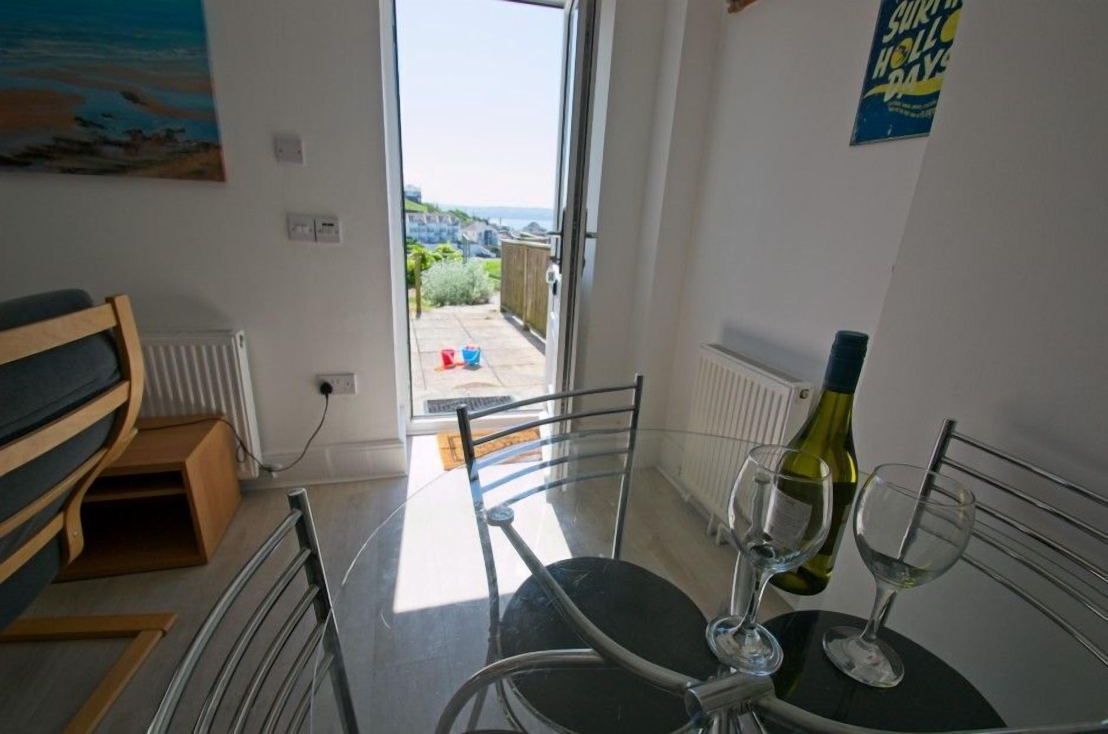 A room with a dining table, chairs, a bench, a bottle of wine, and a glass. There is a view of a garden and a building outside the window.