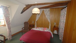 A bedroom with a wooden ceiling, a bed with a red bedspread, a chair, a nightstand with a lamp, and floral curtains. - --Standard room-- (launay blot)