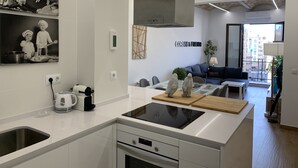 A modern kitchen with white cabinetry, a built-in oven, and a black induction cooktop. There's a dining area with a glass table and chairs, and a living room with a sofa and a large window. - Corsini & Friends a paradise in the center of Tarragona (Tarragona)