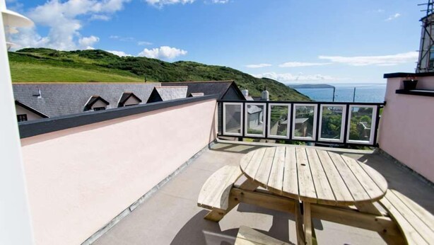 Eine Dachterrasse mit einem Holztisch und Stühlen, Blick auf das Meer und eine grüne Hügelkette im Hintergrund.