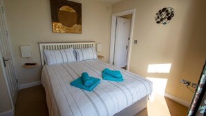 A bedroom with a bed, two rolled towels, a painting, a door, and a wall-mounted light fixture. - Gorgeous 2 bed apartment with large terrace overlooking Combesgate Beach (Woolacombe)