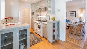 A modern kitchen with white cabinets, a stove, and a microwave. There is a dining area with a wicker sofa and a lamp. - Refined Coastal Escape a Block from West Beach! (Santa Barbara)