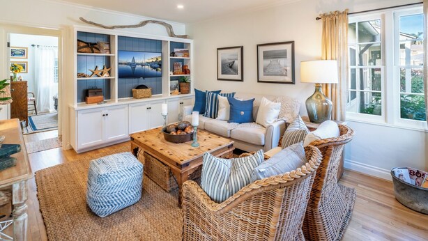 A cozy living room with a sofa set, a coffee table, wicker chairs, a built-in shelving unit with decorative items, and a large window with curtains. - Refined Coastal Escape a Block from West Beach! (Santa Barbara)
