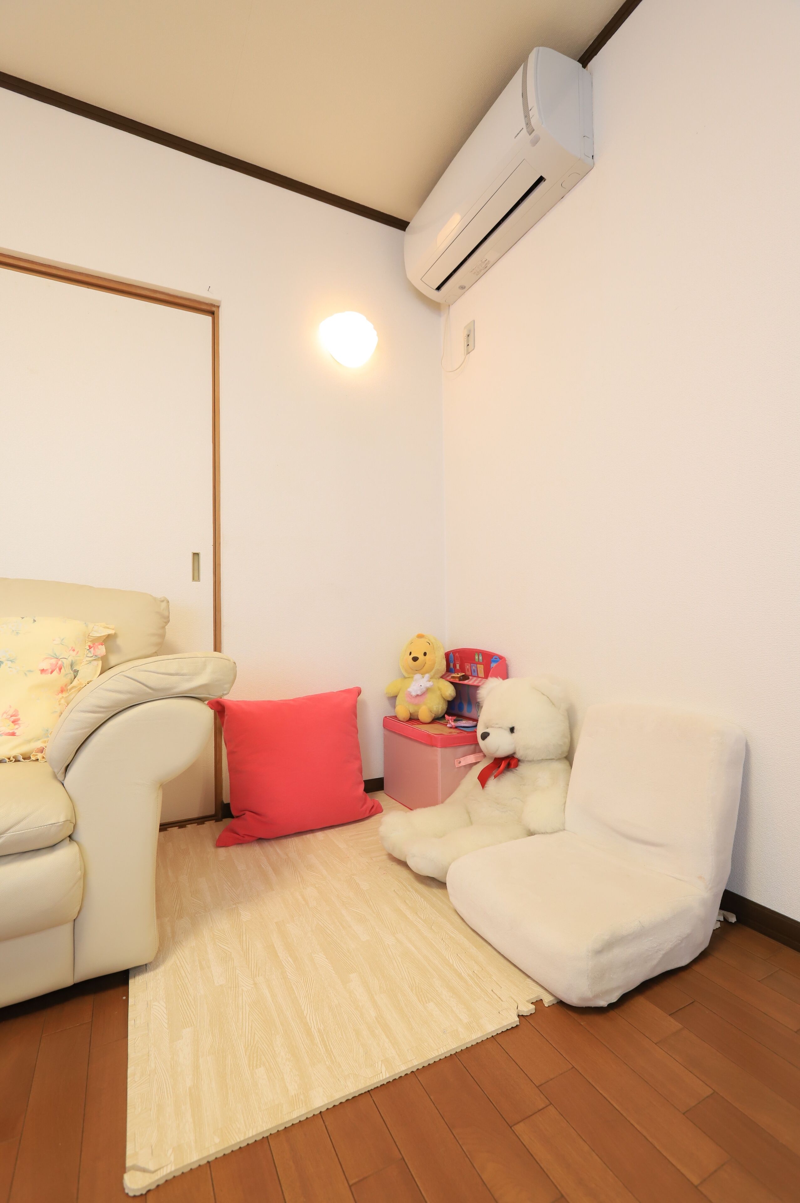 Un ambiente accogliente con un divano color beige, un cuscino rosso, una sedia bianca e un orsetto su un tavolino. Un condizionatore d'aria è fissato al muro.