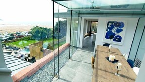 Una habitación moderna con una pared de vidrio con vista a una playa, una mesa de comedor de madera y un cuadro abstracto enmarcado.