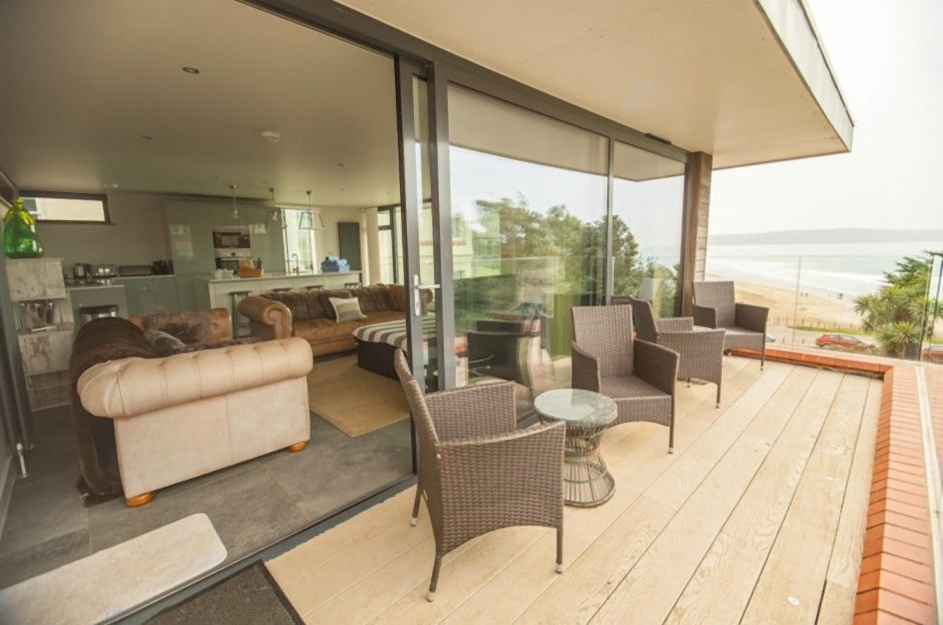 Uma sala de estar moderna com sofá, cadeiras e uma porta de vidro que dá para uma varanda com vista para a praia.