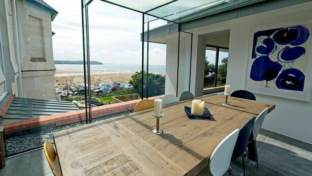 Un comedor moderno con pared de vidrio que da a una playa, una mesa de madera y un cuadro en la pared.