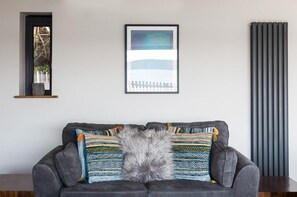 A grey sofa with patterned cushions and a fluffy grey throw, against a white wall with a framed picture and a radiator.
