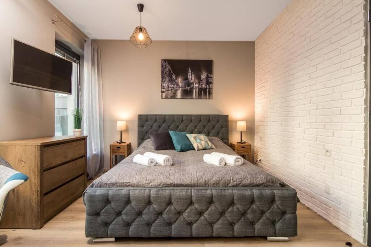 A bedroom with a large bed, two bedside tables with lamps, a wooden dresser, a TV mounted on the wall, and a cityscape painting above the bed.