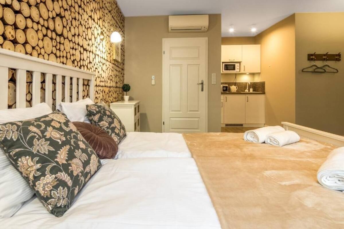 A bedroom with a bed, pillows, a bedside table, a wall-mounted lamp, a door, and a kitchenette in the background.