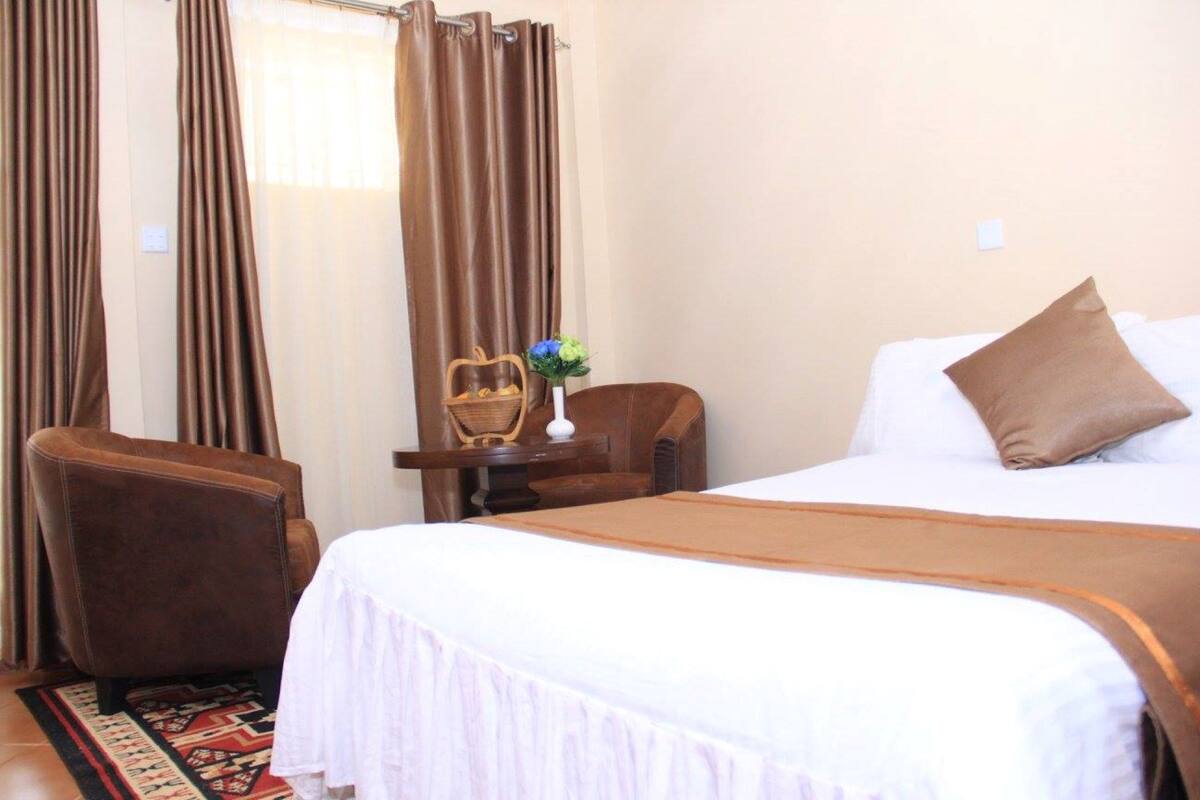 Una habitación de hotel con una cama, una silla, una mesita con un jarrón y cortinas.