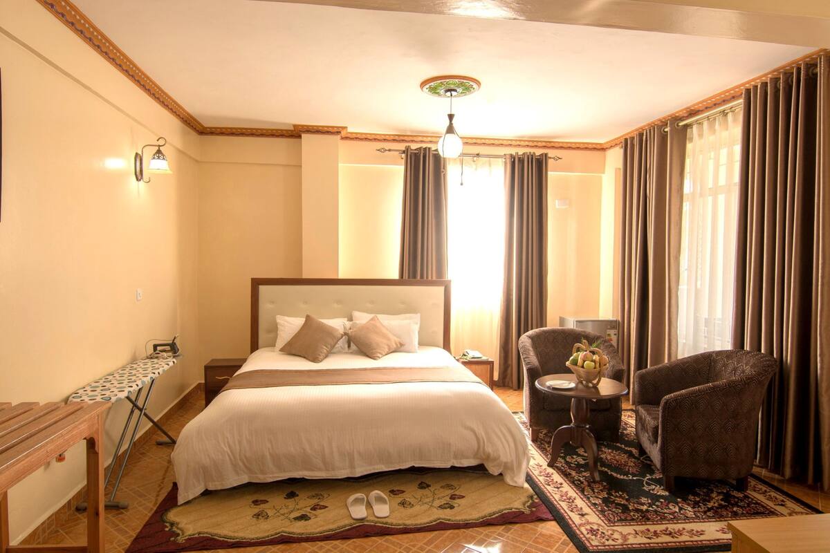 Habitación de hotel con una cama grande, una mesita de noche de madera, una pequeña mesa con lámpara, una silla y una alfombra decorativa.