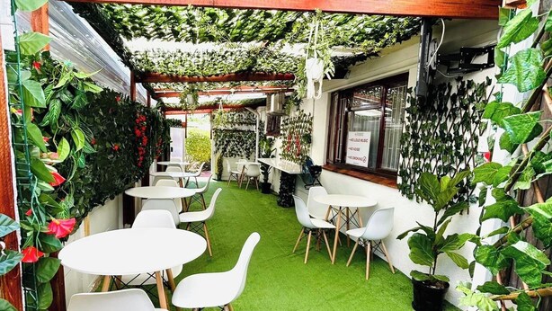 Ein Patio mit Kunstgrasanlage, weißen Tischen und Stühlen, umgeben von üppigen, grünen Pflanzen und Ranken.