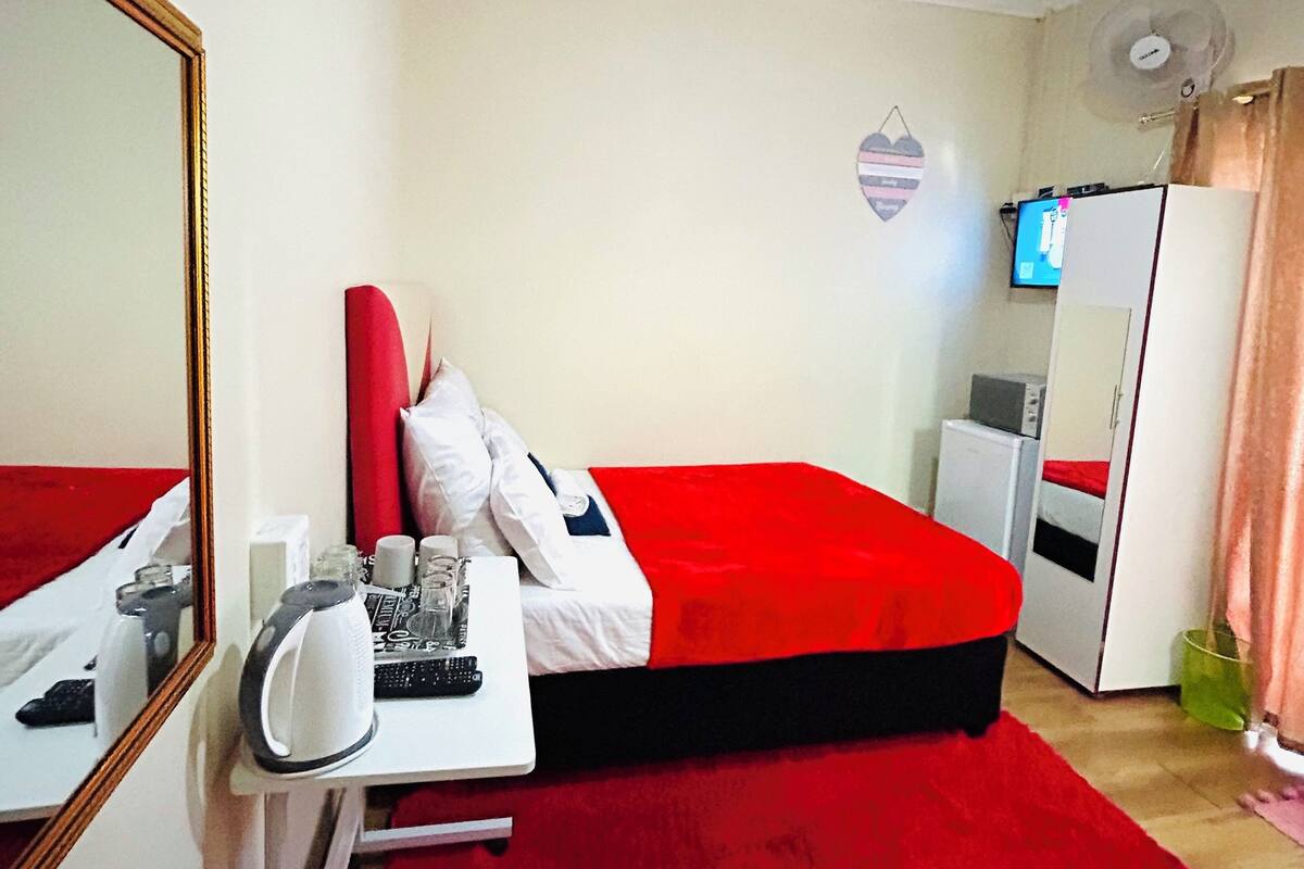 Una habitación de hotel compacta con cama, escritorio, espejo, hervidor y televisor.