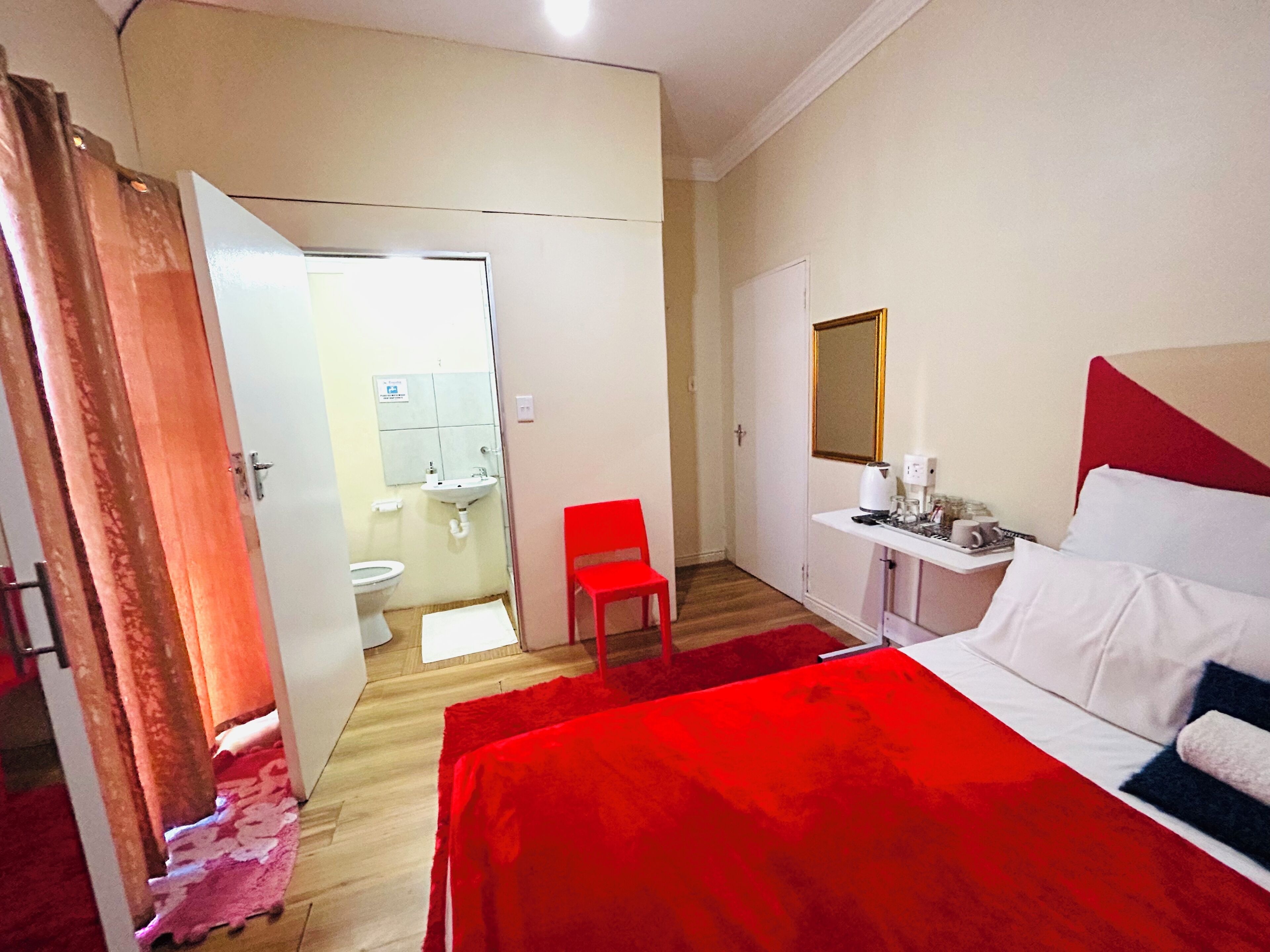 A hotel room with a bed, a desk with a mirror, a red chair, and a bathroom visible through an open door.