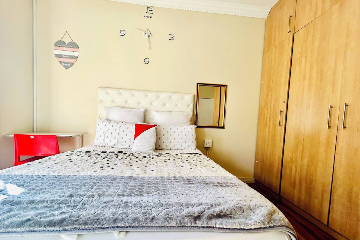 Un dormitorio con una cama, una silla roja, un armario de madera, un reloj de pared y un espejo.