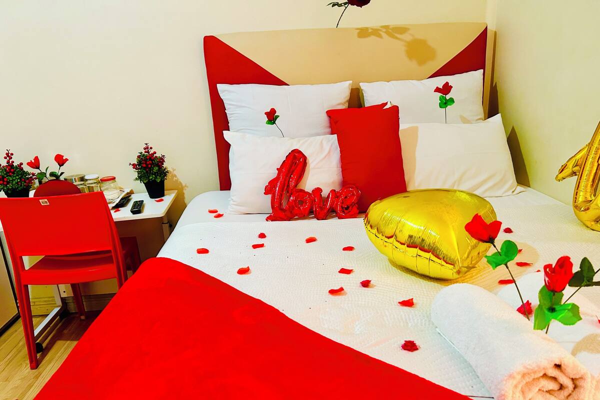 Una habitación de hotel con una cama, una silla roja, una mesita con una planta y una pared decorada con una rosa roja.
