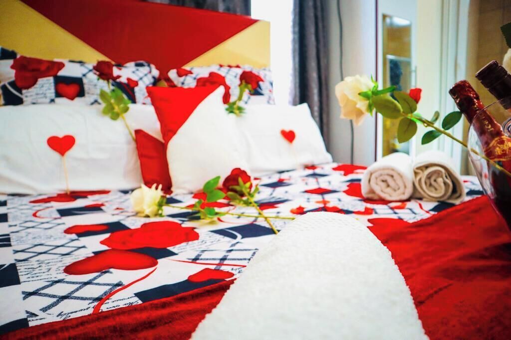 A hotel room with a bed featuring a red and white quilt with heart patterns, white pillows, and a bouquet of roses.