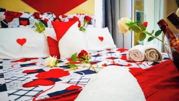 Quarto de hotel com cama coberta por um edredom vermelho e branco com estampas de corações, almofadas brancas e um buquê de rosas.