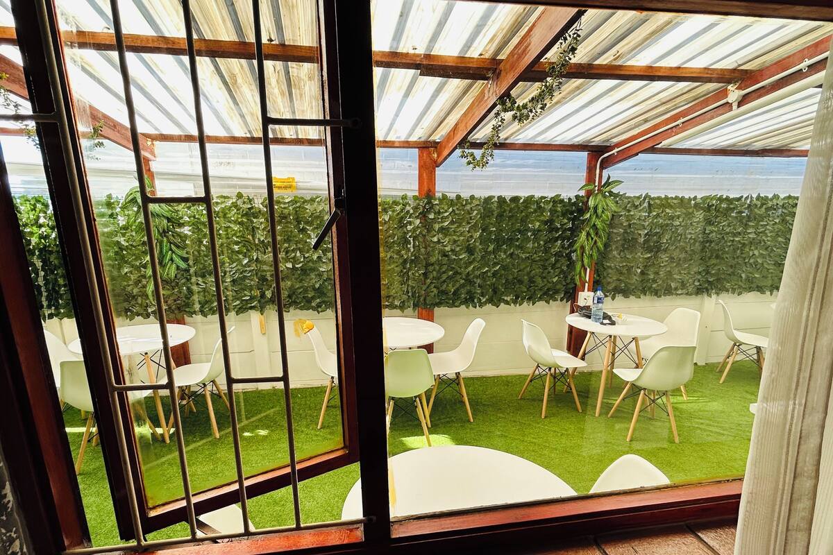Un patio cubierto con mesas y sillas blancas redondas, césped artificial verde y vistas a un jardín a través de una ventana con marco de metal.