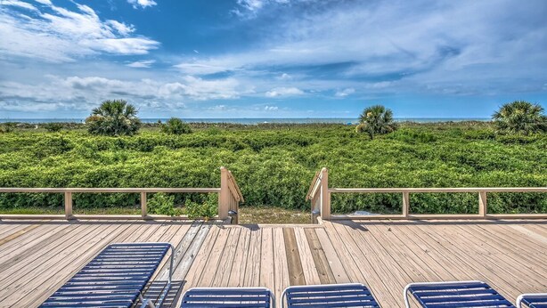 A wooden deck with blue lounge chairs facing a lush green landscape under a clear sky.
