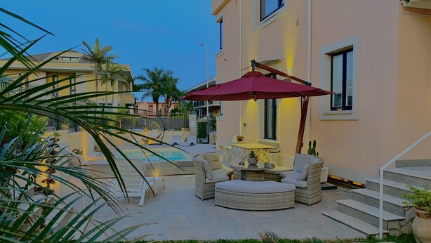 A pool area with lounge chairs and a red umbrella, surrounded by greenery and a building with a light-colored facade. - HOLIDAY HOME "U SCRUSCIU RO MARI" (Avola)