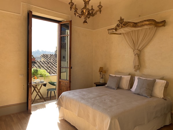 A bedroom with a large bed, a chandelier, a nightstand with a lamp, and a view of the outside through an open door.