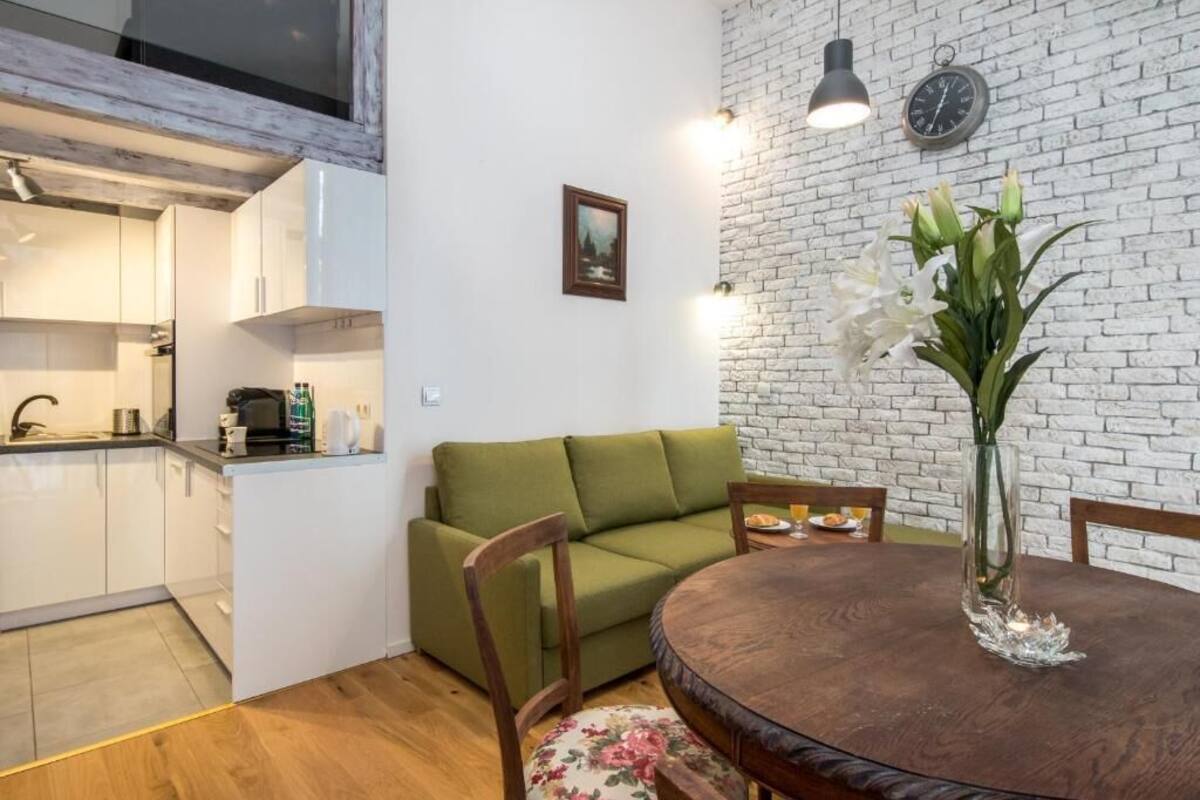 A compact living space with a kitchen, a green sofa, a dining table with chairs, and a vase with flowers.