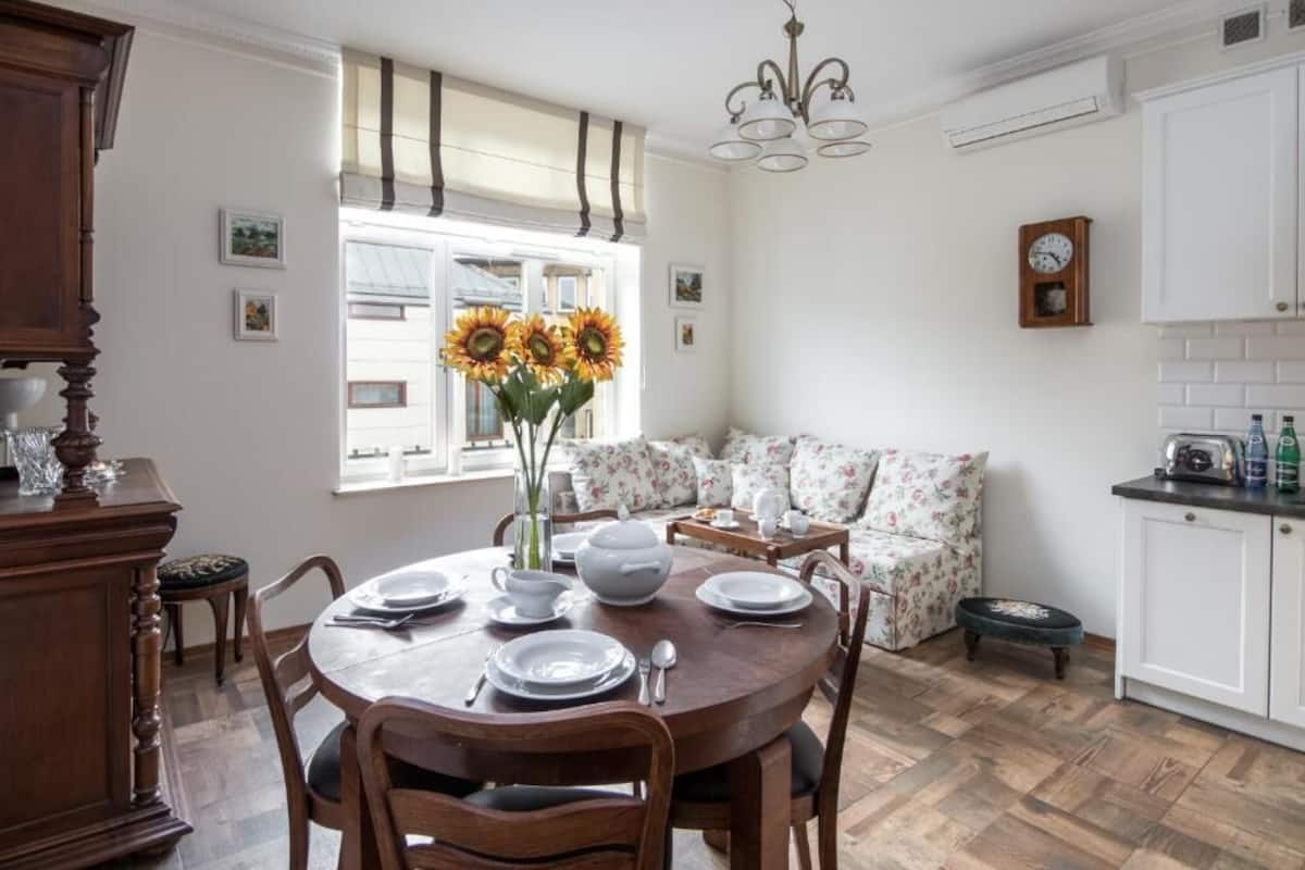 A dining area with a round wooden table set for four, surrounded by wooden chairs. There is a floral sofa with a small table beside it, and a window with curtains and sunflowers on the sill. A wooden cabinet and a small stool are also visible.