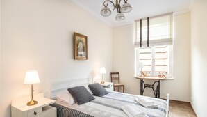 A neatly arranged bedroom with a bed, bedside lamps, a small table, and a window with shutters. - Apartments Wygoda A4You (Kraków)