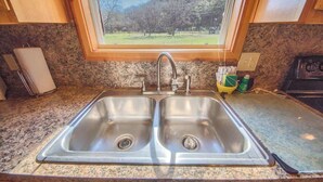 A kitchen with a double stainless steel sink, granite countertop, and a window overlooking a grassy area. - RR 9122WC Wine Cup Cabin (New Braunfels)