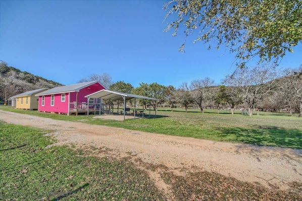 Um edifício rosa com varanda coberta, cercado por um campo gramado e árvores.