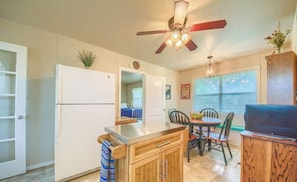 Une cuisine équipée d’un réfrigérateur, de meubles de rangement en bois, d’une table à manger avec des chaises, d’un ventilateur de plafond et d’une fenêtre avec des stores.