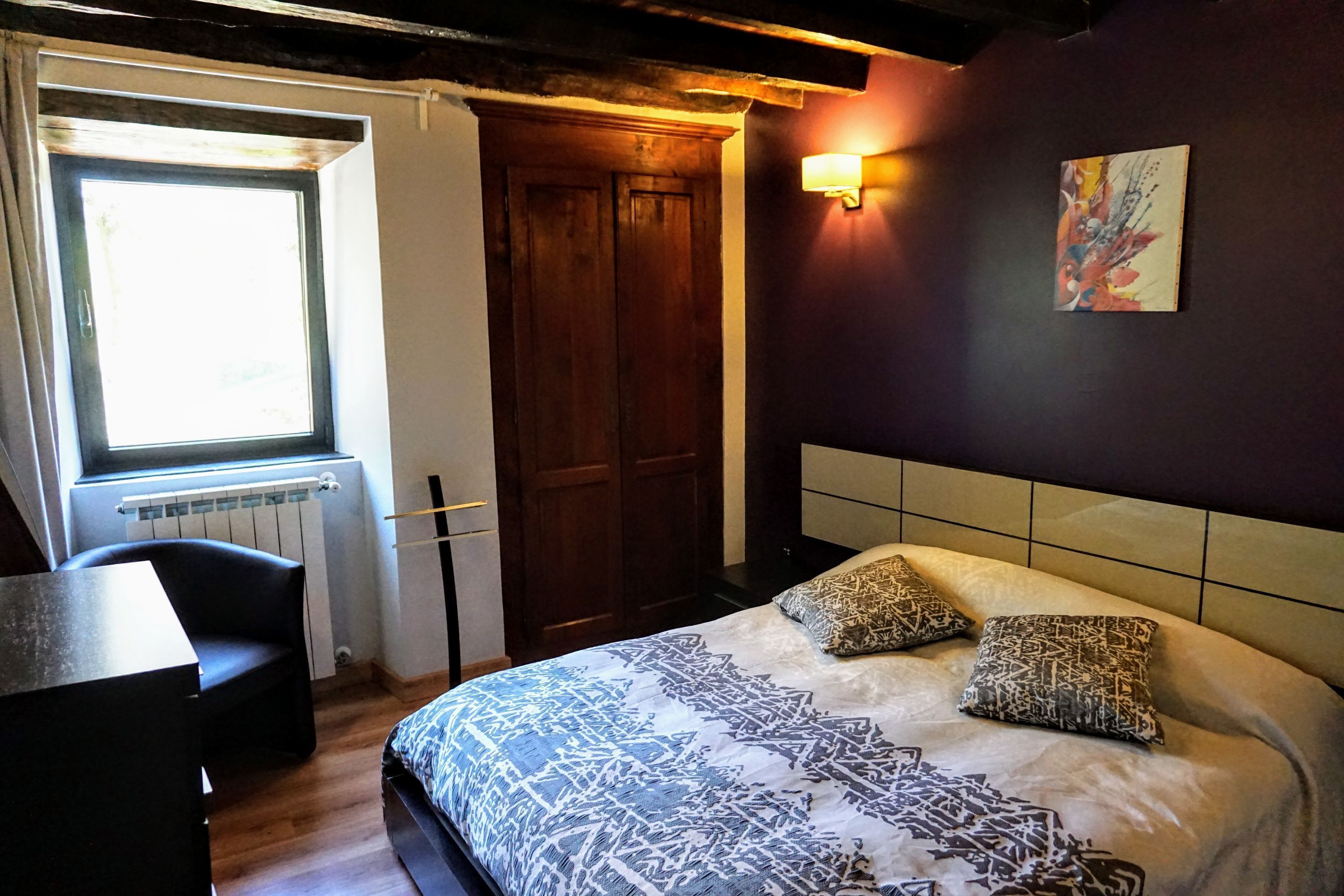 A bedroom with a bed, a chair, a window, a radiator, and a wall-mounted light fixture.