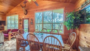 L’intérieur d’un chalet en bois comprend une table à manger, des chaises, un ventilateur de plafond et une grande fenêtre donnant sur une forêt.