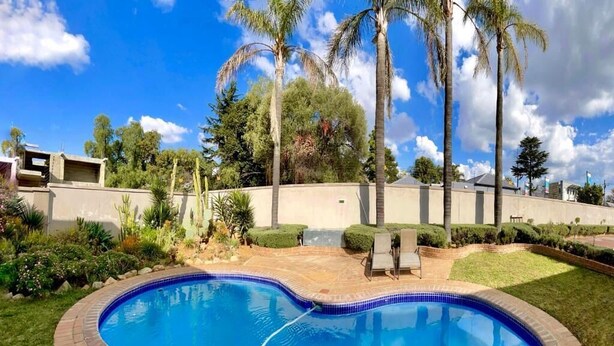A swimming pool with a tiled edge, surrounded by a brick border, palm trees, and a well-maintained lawn. - Azinnia Rivonia Guest House (Sandton)