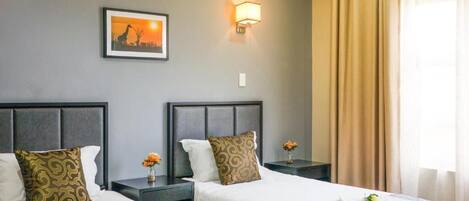 Quarto de hotel com duas camas, uma mesa de cabeceira com abajur, um quadro na parede e uma janela com cortinas.