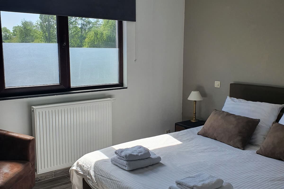 Un dormitorio con cama, una silla, una mesita de noche, una lámpara y una ventana que ofrece vistas a árboles y agua.