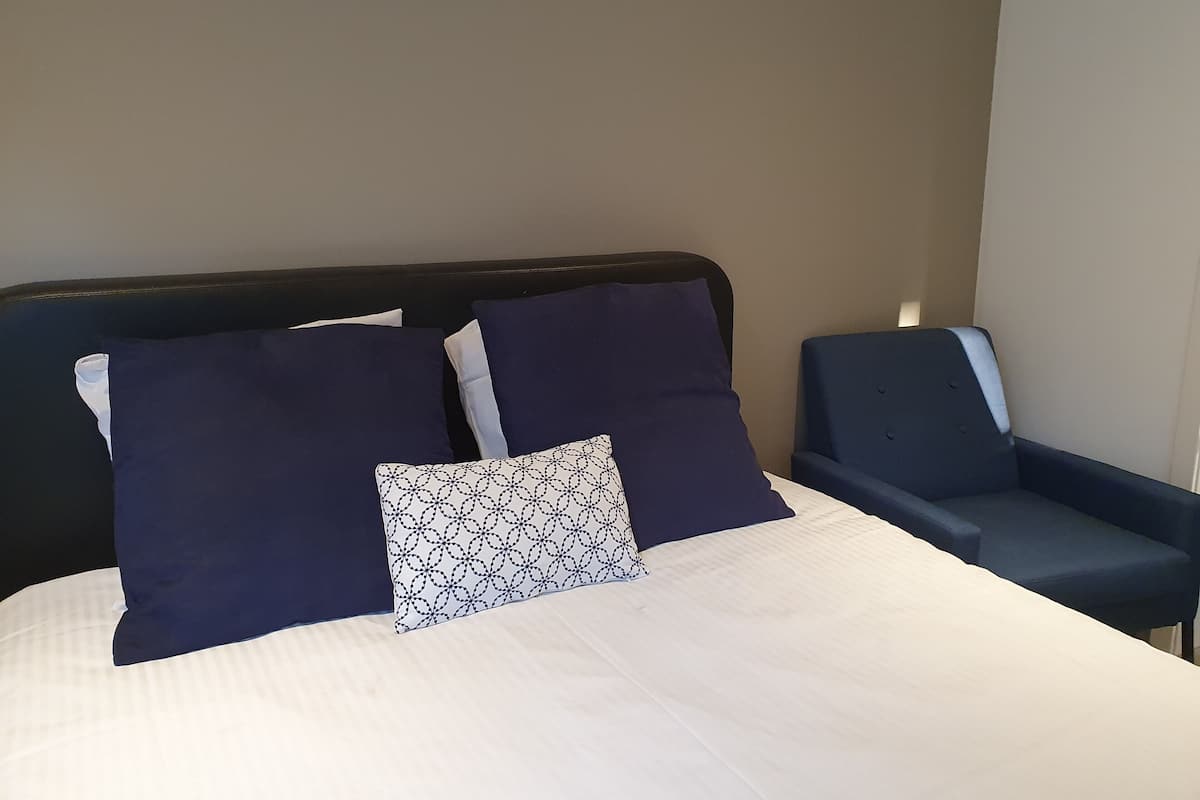 Una cama bien hecha con almohadas azul oscuro y blancas, un cojín estampado y un sillón azul a su lado.