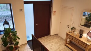 A room with a wooden floor, a dark wooden door, a small wooden desk with drawers, a mirror, and a potted plant. - Memories (Ghent)