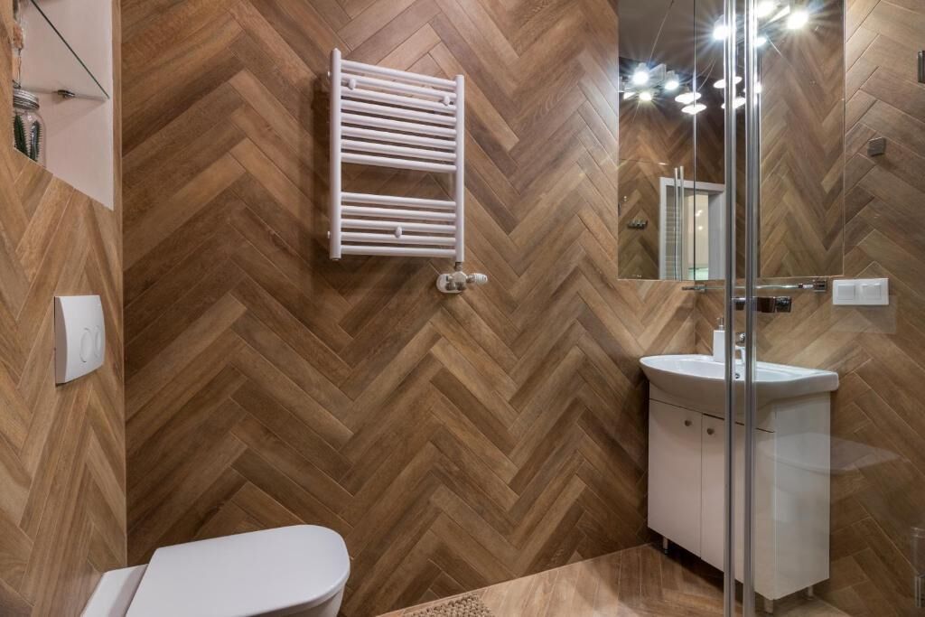 A bathroom with a herringbone wood floor, a white toilet, a glass shower enclosure, and a white sink with a mirror above it.