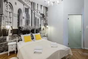 Quarto de hotel com um grande mural de uma rua da cidade, cama com roupa de cama branca e almofadas amarelas, mesa de cabeceira e um candelabro.