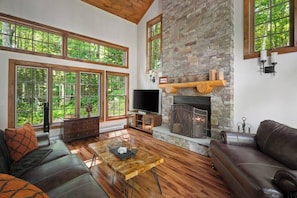 A spacious living room with a stone fireplace, large windows, and a television.