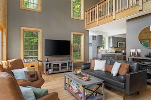 A spacious living area with a high ceiling, a television, a foosball table, and a kitchen in the background.