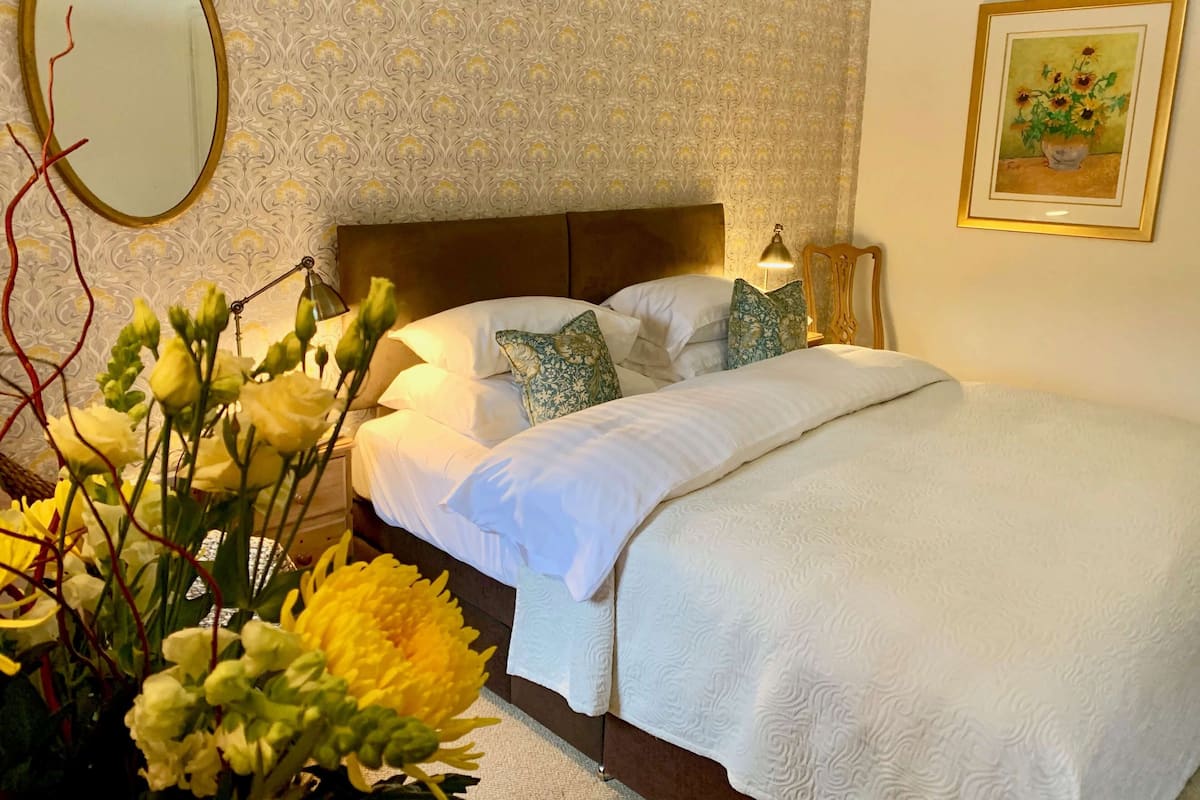 Un lit bien fait, avec du linge de lit blanc, une tête de lit et des coussins décoratifs. Une table de chevet avec une lampe et un tableau encadré ornent le mur.