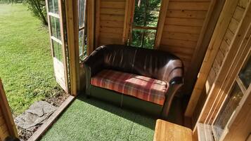Una cabaña de madera con suelo de césped artificial verde, un sofá de cuero marrón con un cojín escocés y un banco de madera.